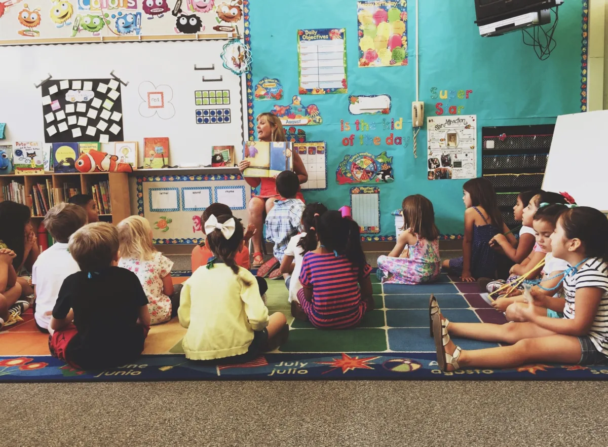 An image of a teacher reading a book to her classroom while her students are sitting in the floor.