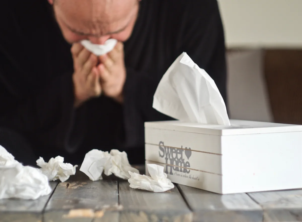 An image of a future male vybe urgent care patient blowing his nose in front of a coffee table filled with used tissues and a tissue box.