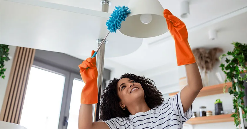 An image of a young housekeeper in apron dusting a lamp in the living room.