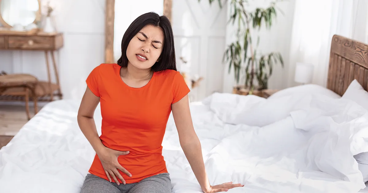 An image of a woman sitting on the side of her bed in pain due to having a UTI.