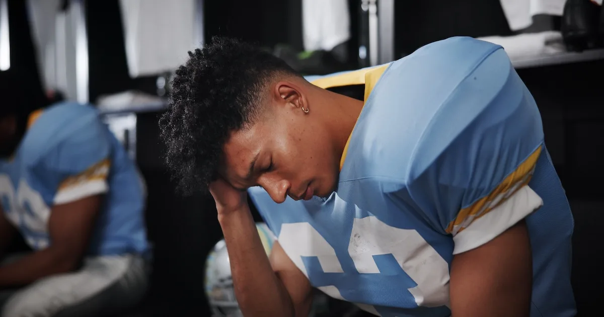 A young football player in a blue and yellow jersey sits in a locker room holding his head, showing signs of a concussion.