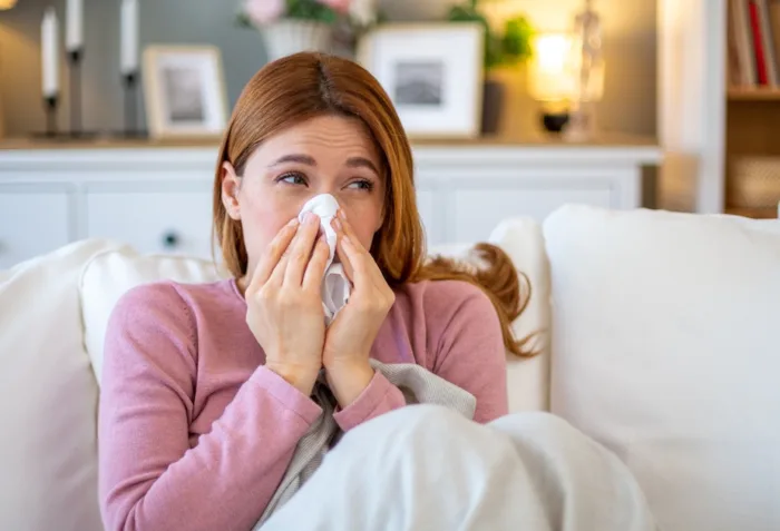 A woman in a pink sweater rests under a blanket on a couch, holding a mug and a tissue to her head while sick with a cold.