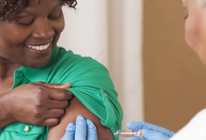 A smiling Black woman in a green shirt prepares to get a flu shot in her arm from a healthcare worker in blue gloves.