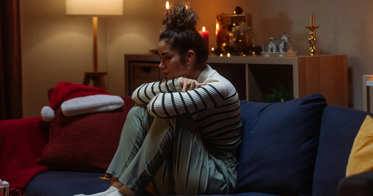 A woman sits on a couch hugging her knees, experiencing post-holiday burnout in a room with festive lights and a Santa hat.