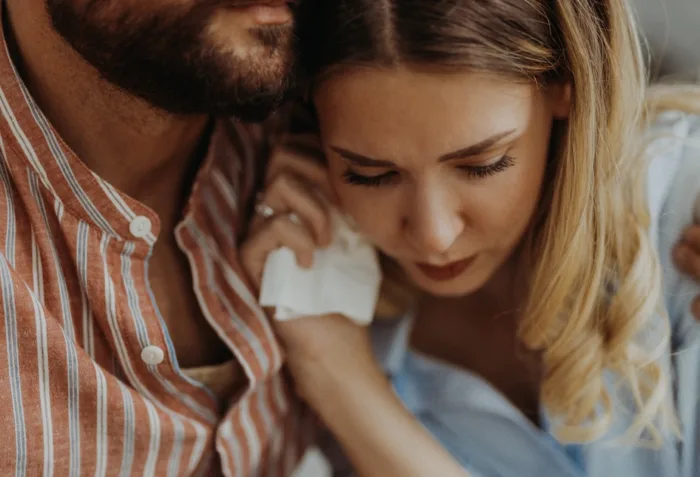 A bearded man in a striped shirt embraces a blonde woman holding a tissue as they discuss untreated STDs and fertility.